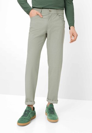 Mann trägt hellgraue Hosen, die an den Knöcheln hochgekrempelt sind, ein grünes Langarmshirt und grüne Wildledersneaker mit Gummisohlen.