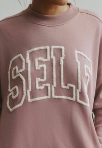 Roosa roosakas dressipluus soonikkoelusega kaeluse ümber, millel on suured, tekstuuriga valged tähed "SELF". Pehme kangas sileda tekstuuriga.