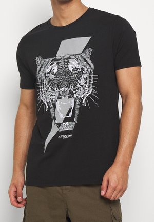 Homme portant un t-shirt noir avec un grand motif de tigre rugissant et un design d’éclair gris sur le devant, associé à un pantalon marron.