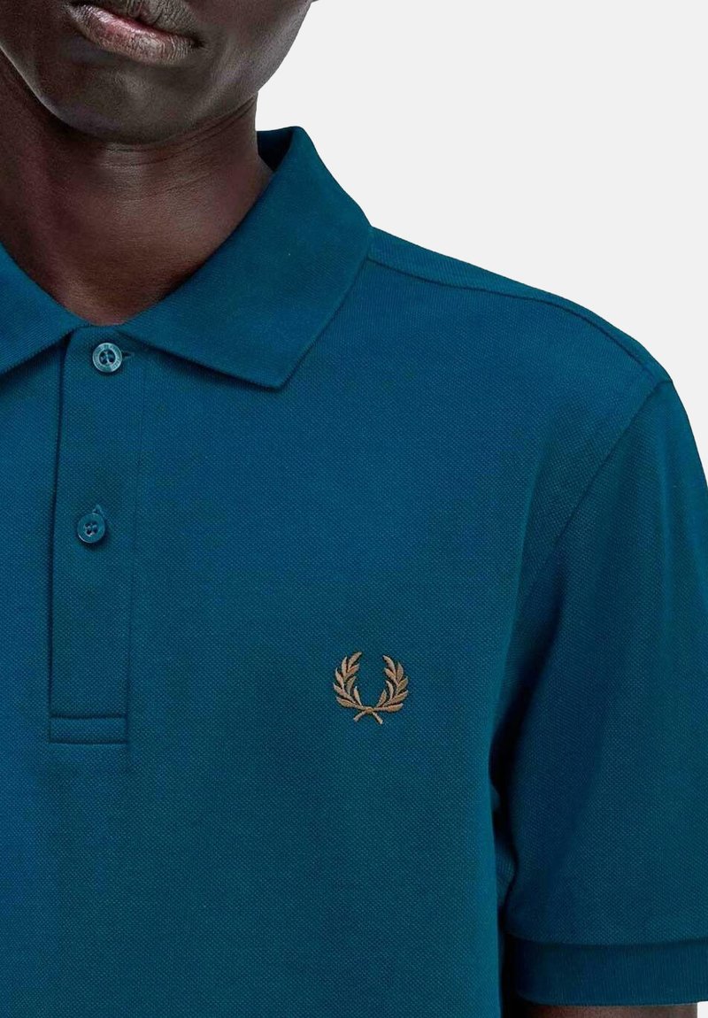 Polo shirt di un verde scuro con un tessuto texturizzato, due bottoni e un piccolo logo d'alloro dorato nell'area del petto sinistro. Maniche corte, colletto classico.