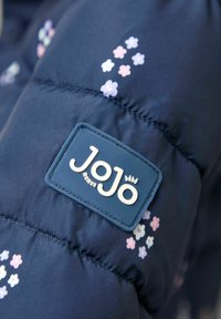 Tumesinine pufi jakk, millel on roosa ja valge lillemuster. Omab ristkülikukujulist brändi sildiga, millel on tekst "JoJo maman bébé".