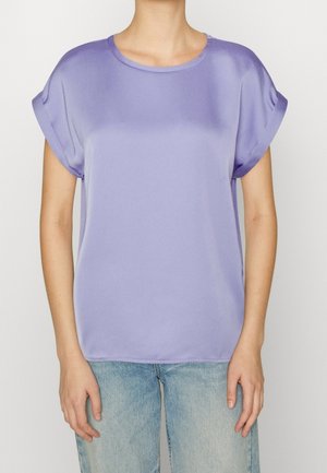 Femme portant un chemisier en satin lavande à manches courtes avec des poignets retroussés et un jean bleu clair, debout devant un fond uni.