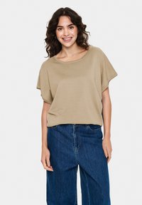 Beige, rövid ujjú felső széles nyakkivágással és laza szabással, sötétkék, bő szabású farmernadrággal párosítva, amely egy laza és kényelmes megjelenést mutat.