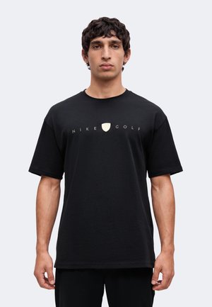 Mladý muž má na sebe čierne tričko Nike Golf s malým štítovým logom a textom na hrudi, stojí pred jednofarebným pozadím.