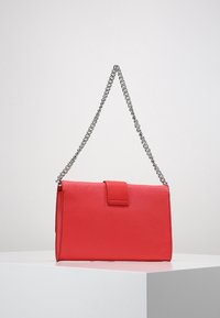 Sac à main en simili cuir rouge avec fermeture à rabat, surface texturée et bandoulière en chaîne argentée, présentant une forme rectangulaire et un design minimaliste.