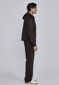 Bruine hoodie met ritssluiting en een gevoerde capuchon, gecombineerd met bijpassende taps toelopende broek. Zachte stof, relaxte pasvorm en witte sneakers met beige zolen.