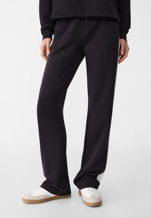 Personne portant un pantalon noir ample et des baskets blanches, debout devant un fond clair uni.