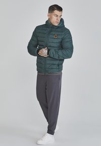 Mörkgrön puffjacka med huva och dragkedja, med en liten logotyp. Bärs med grå byxor och vita sneakers, står i en neutral pose.