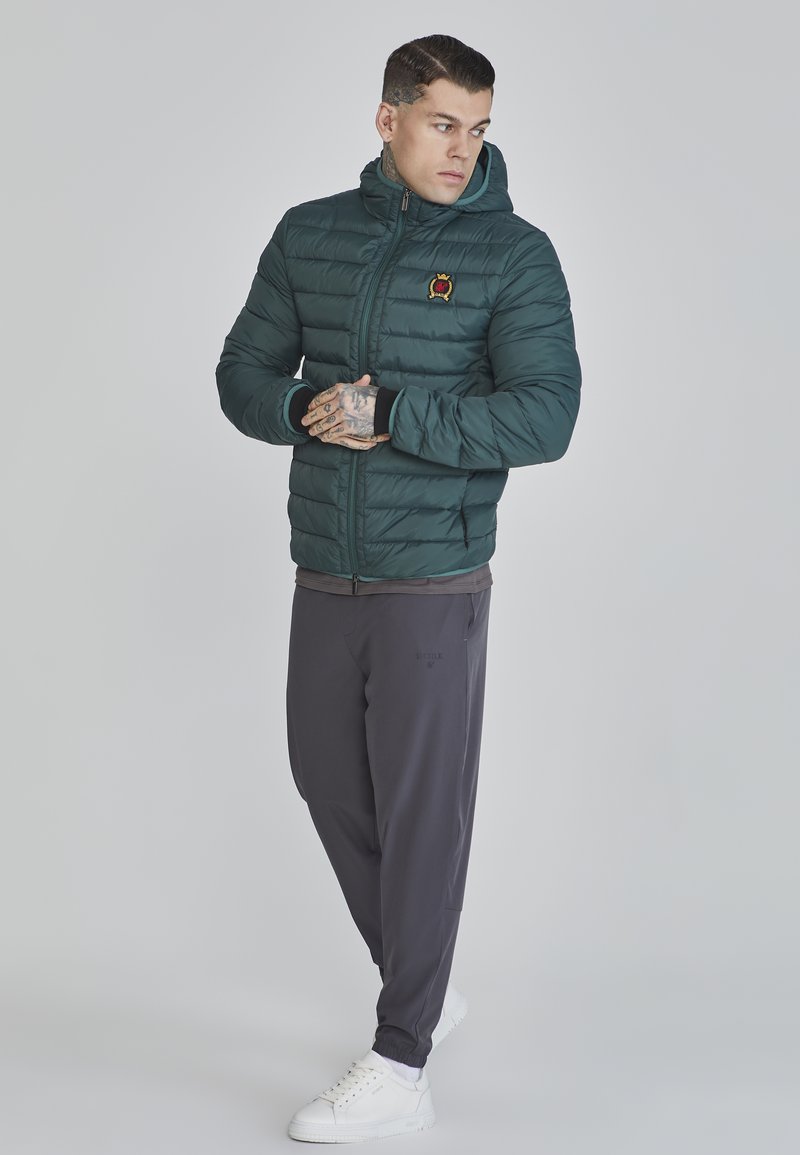 Giacca a vento verdi scuro con cappuccio e zip, caratterizzata da un piccolo logo. Indossata con pantaloni grigi e sneakers bianche, in una posa neutra.