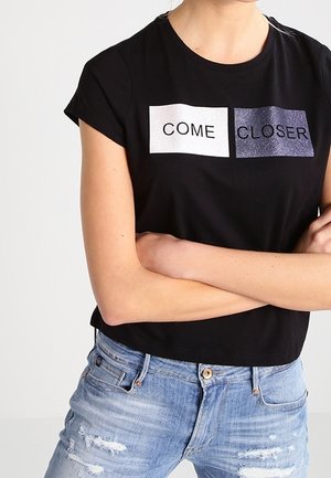 Person som bär en svart t-shirt med texten "KOM NÄRMARE" i vitt och glittriga svarta block, tillsammans med slitna ljusblå jeans.