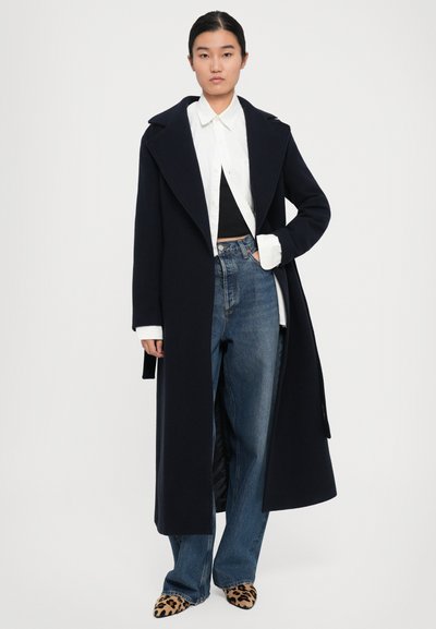 Donna che indossa un lungo cappotto scuro su una camicia bianca, una maglietta nera, jeans blu e scarpe a punta con stampa leopardata, in piedi su uno sfondo bianco.