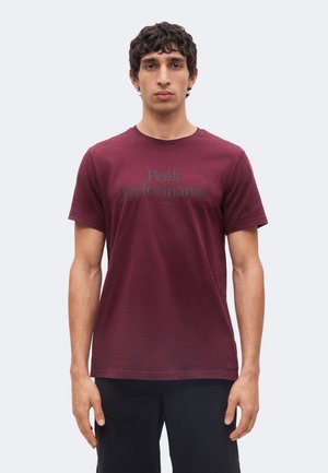 Jeune homme aux cheveux foncés et bouclés portant un t-shirt uni bordeaux à manches courtes avec l'inscription « "Peak Performance" » en lettres foncées.