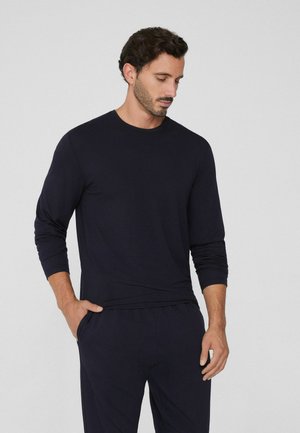Homme avec des cheveux courts et bouclés et une barbe, portant une chemise noire à manches longues et un pantalon noir, se tenant avec une main dans la poche, regardant vers le bas.