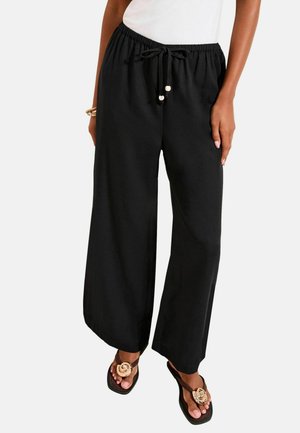 Woman wearing loose black drawstring pants, white top, gold bracelets, and black sandals with large circular gold decorations.