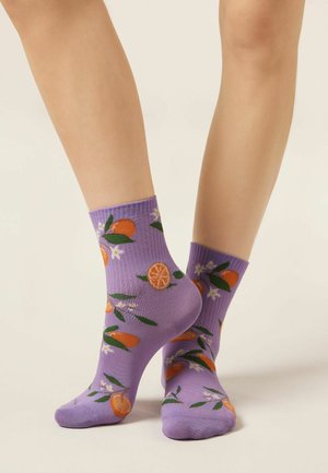 Gambe con calzini viola fino alla caviglia con motivi di frutti agrumi arancioni e verdi e fiori bianchi su uno sfondo chiaro uniforme.