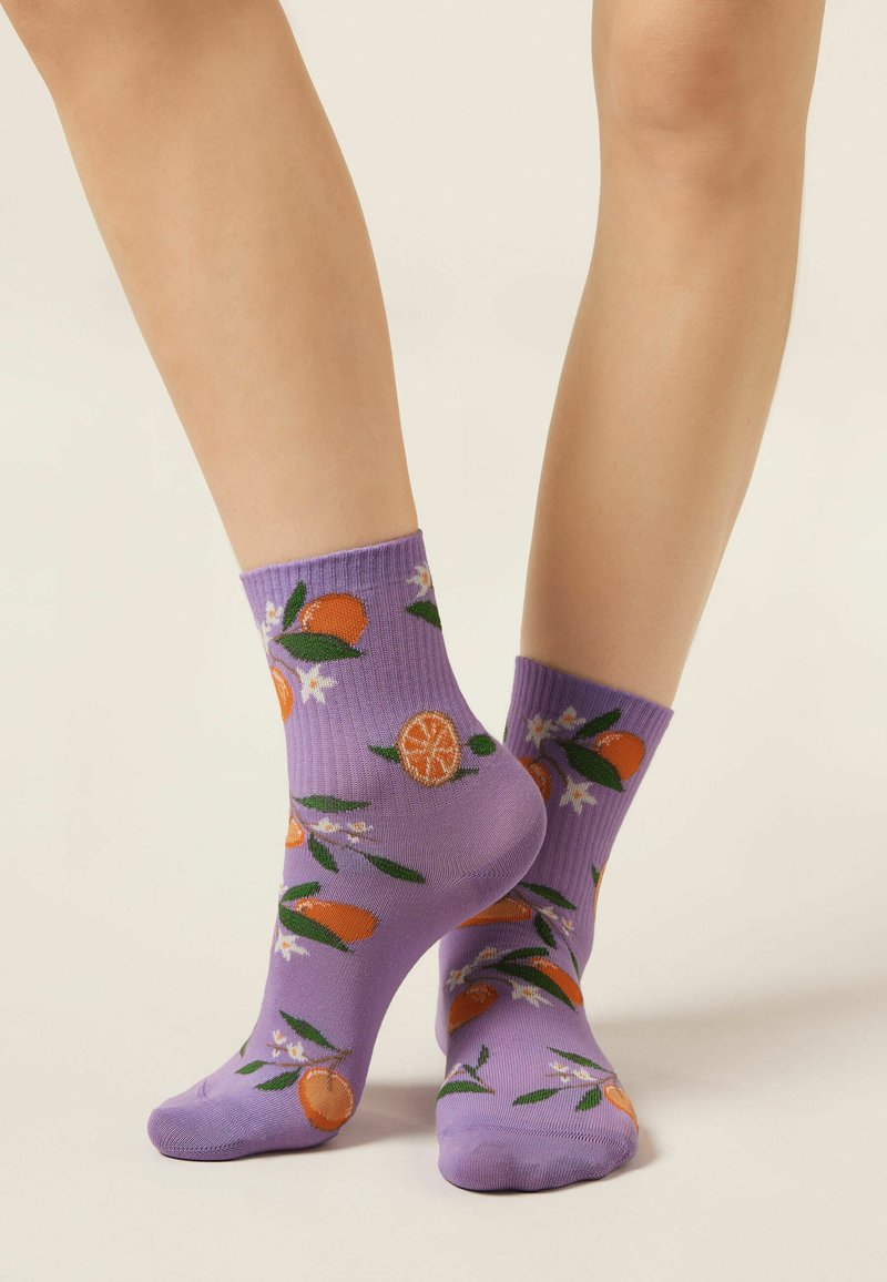 Jambes portant des chaussettes courtes violettes ornées de motifs d'agrumes orange et verts ainsi que de fleurs blanches sur un fond clair uni.