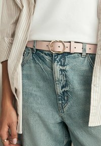 Cintura in pelle rosa chiaro con una fibbia circolare dorata, indossata sopra un paio di jeans in denim azzurro chiaro strappati. Camicia a righe visibile sopra la cintura.