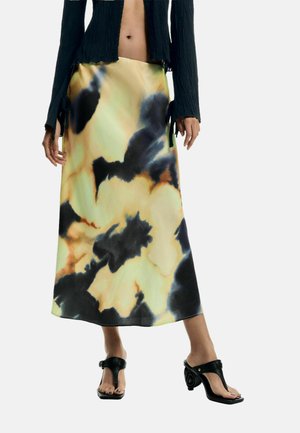 Naine kannab tie-dye mustriga midi-seelikut, millel on mustad, beežid ja oranžid mustrid, musti avatud varbaga kontsakingi ja kärbitud musta pikema varrukaga t-särki.