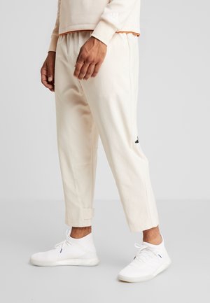 Man draagt lichtbeige broek, witte gebreide sneakers en een beige Adidas-trui, staand tegen een effen lichtgrijze achtergrond.