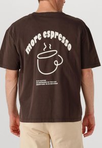 T-shirt in cotone marrone con testo bianco sul retro: "more espresso", accompagnato da un'illustrazione di una tazza di caffè e dettagli di preparazione sottostanti.