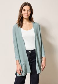 Ljus teal kavaj med långa ärmar, två framfickor, öppen framsida och en texturerad yta, bärs över en vit topp och mörka jeans.