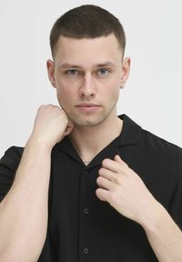 Musta lyhythihainen paita, jossa on kaulus ja napitusyksityiskohtia. Mallilla on vaalea iho, lyhyt hiustyyli ja hopeinen renkaanikorvake.