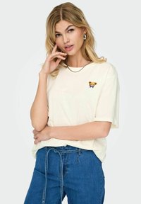 T-shirt en coton crème avec un petit ours brodé portant un nœud bleu sur la poitrine. Associé à un jean en denim bleu. Manches courtes, coupe décontractée.