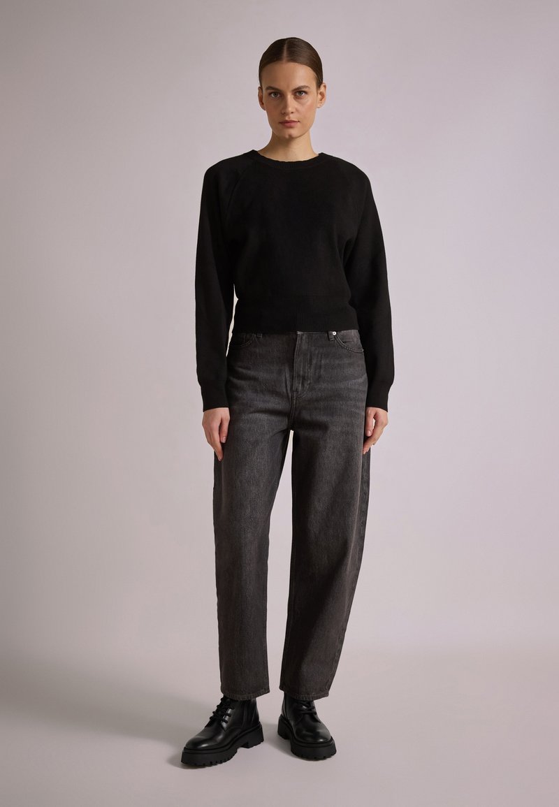 Black sweater with a ribbed hem paired with high-waisted dark grey jeans. Model wears black lace-up boots, standing against a neutral background.