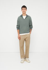 Pulover verde cu dungi, cu guler tip șal, asortat cu pantaloni bej și adidași albi. Modelul stă în fața unui fundal alb simplu.