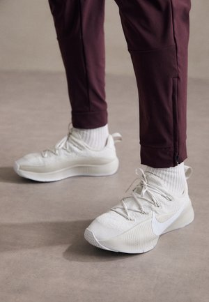 Ténis de desporto brancos com superfície texturizada, apresentando detalhes cinzentos, atacadores planos e logótipo da Nike, combinados com calças de desporto bordeaux e meias brancas.