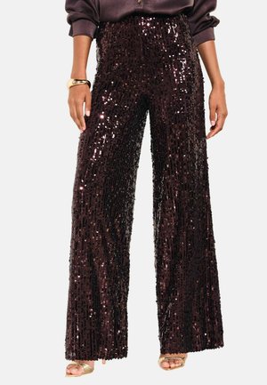 Woman wearing high-waist wide-leg black sequin pants with gold open-toe heels and a dark long-sleeve top, showing hands relaxed by sides.