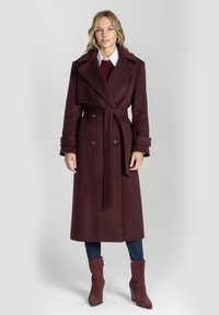 Bordowy długi płaszcz z podwójnym zapięciem, szerokimi klapami i wiązaniem w talii. Miękka tkanina o gładkiej fakturze, zestawiona z dopasowanymi dżinsami i botkami za kostkę.