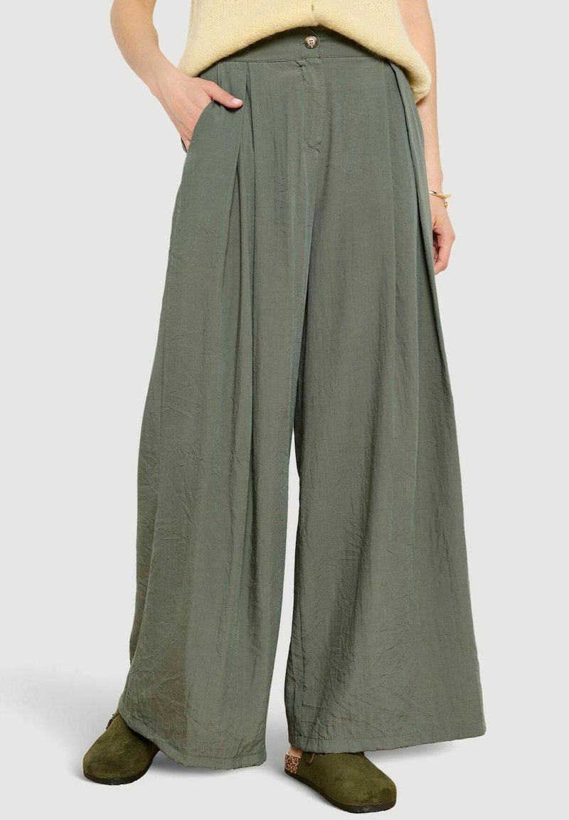 Inimene, kes kannab laiade jalgadega oliivrohelisi pükse, helekollast kampsunit ja oliivirohelisi sussilaadseid kingi, ühe käega taskus ja randmel käevõrud.