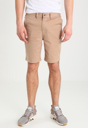 Mężczyzna w beżowych szortach do kolan, białej koszulce i szarych butach sportowych, stojący na tle jasnego, jednolitego tła.