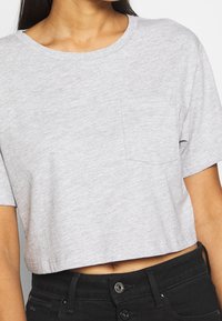 Kvinna i en ljusgrå kort t-shirt med bröstficka och svarta högmidjade jeans, som visar magen och knappar.