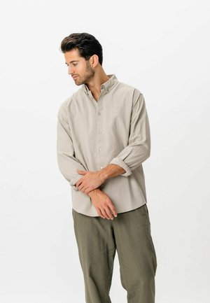 Młody mężczyzna w beżowej koszuli na guziki z podwiniętymi rękawami i oliwkowych spodniach stoi, patrząc w dół i poprawiając zegarek na nadgarstku.