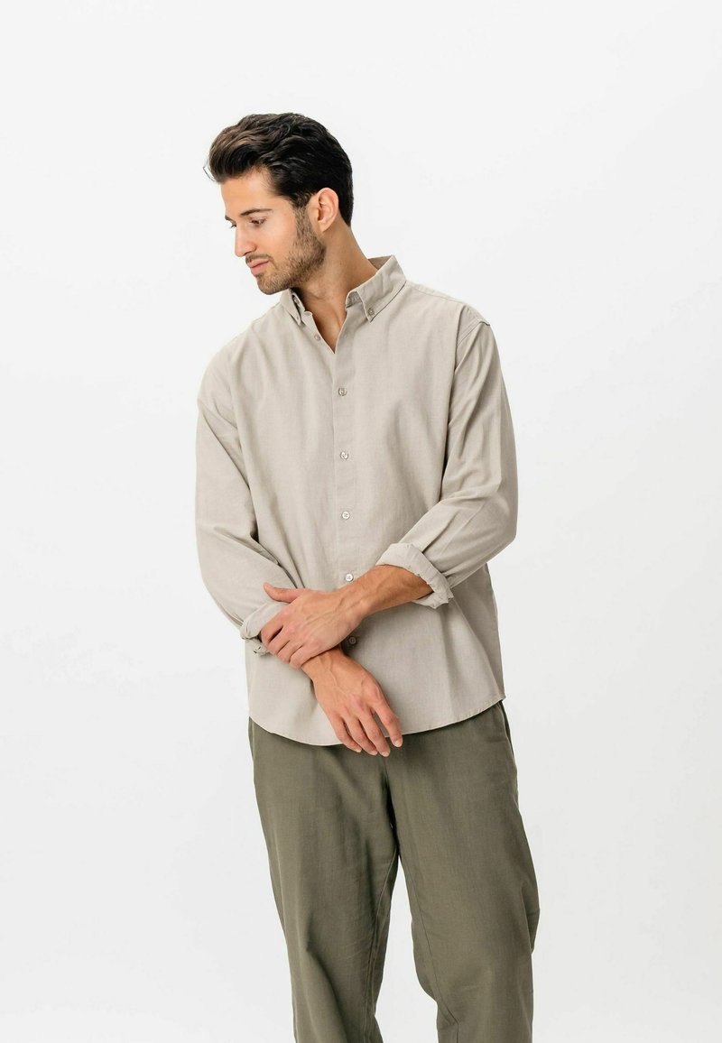 Jeune homme en chemise beige boutonnée aux manches retroussées et pantalon olive, debout, regardant vers le bas et ajustant son poignet.
