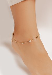 Cavigliera in oro e argento con piccoli pendenti a forma di cuore, posta su un tono di pelle. Texture liscia e design delicato.