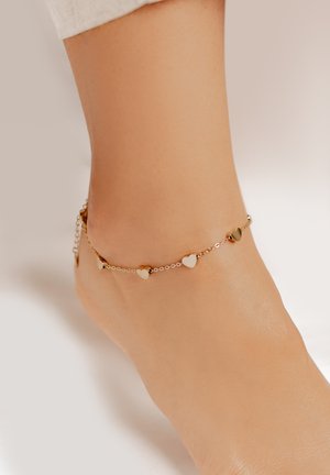 Gold- und Silberkettchen-Anklet mit kleinen herzförmigen Charms, vor einem Hautton platziert. Glatte Textur und zartes Design.