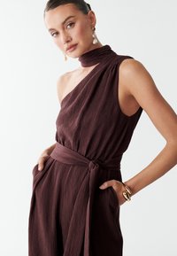 Robe marron texturée à une épaule, avec un lien fin autour du cou, une taille froncée et des poches ; assortie de boucles d'oreilles et d'un bracelet en or et perles.