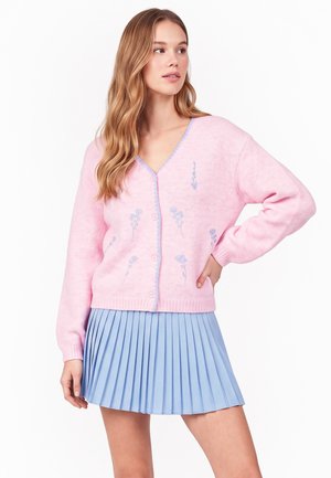 Donna con capelli lunghi che indossa un cardigan rosa floreale con bottoni e una gonna plissettata azzurro chiaro, posando con una mano sui fianchi.
