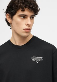 Νέος με κατσαρά μαλλιά που φοράει μαύρο T-shirt με λευκό κείμενο "δημοφιλής προπόνηση: Πάντα αργοπορημένος" και το λογότυπο Puma στο στήθος.