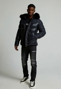 Marineblå vattert jakke med hette trimmed med pels, glidelåslukking og quiltdesign. Brukt med slitte mørke jeans og svarte joggesko.