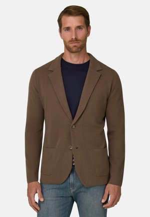 Homme portant un blazer marron à deux boutons sur une chemise bleu marine et un jean bleu, debout devant un fond clair uni.