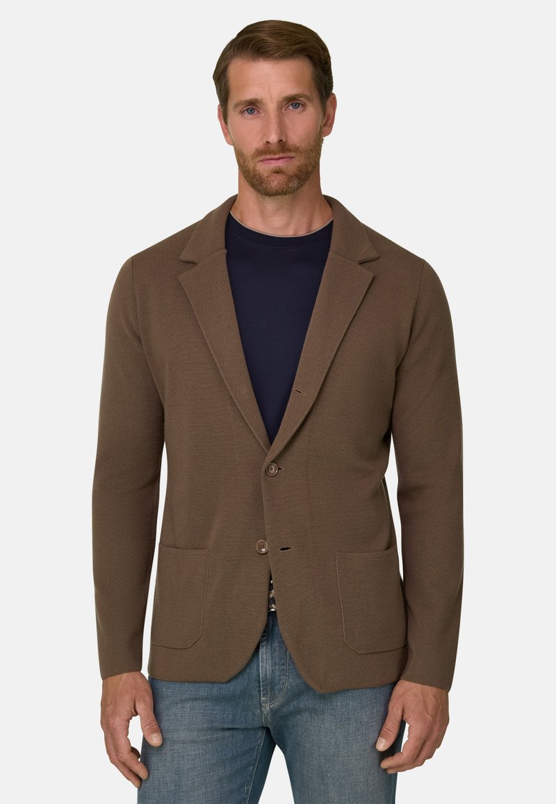 Homme portant un blazer marron à deux boutons sur une chemise bleu marine et un jean bleu, debout devant un fond clair uni.