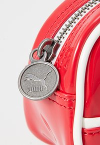 Röd elegant handväska med glänsande yta, med en silverfärgad dragkedja och rund logotagg. Accentuerad med vita detaljer längs kanterna.