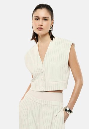 Giovane donna che indossa un gilet corto bianco senza maniche a righe sottili e pantaloni abbinati a vita alta, con orecchini e braccialetto argentati, mani in tasca.