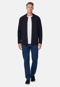 Jūras spēku kardigāns ar apkakli, priekšējiem pogām un divām sānu kabatām, kopā ar baltu krekla un ziliem džinsiem, valkāts ar tumšiem apaviem.