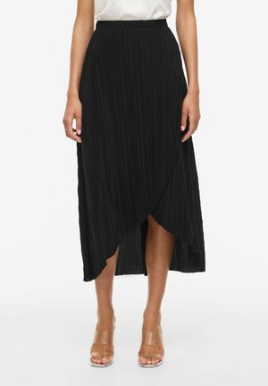 Kvinne iført svart plissert omslagskjørt med asymmetrisk kant, kombinert med høye hæler med klare remmer og hvit topp.