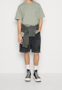 Lichtgroene katoenen t-shirt met een borstzak. Donkergrijze shorts met rafelige randen en zakken. Zwarte hoge sneakers met witte zolen.